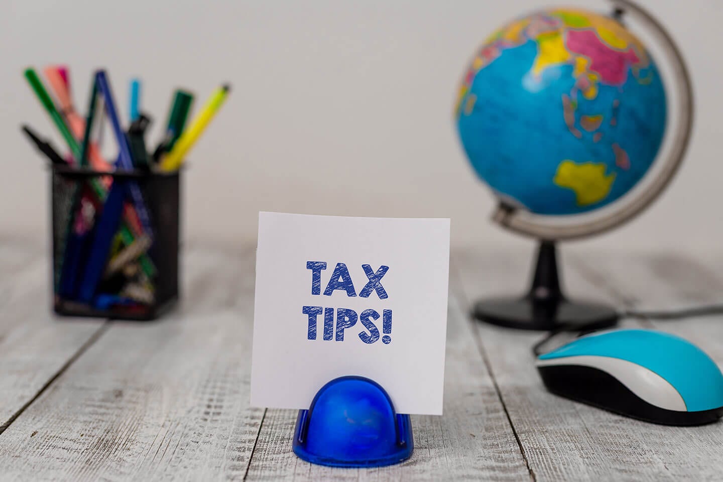 Small globe with colorful pencils and a blue mouse sitting on a wooden desk with a sign that says tax tips