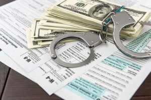 Tax forms typing on a wooden surface with money and handcuffs stacked on top representing tax scam