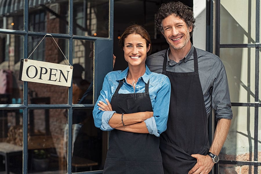 Man and woman in black aprons standing next to a navy-blue door with windows and a business open sign hanging from the door