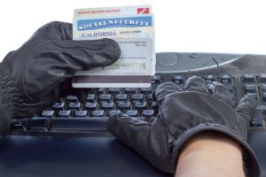 Person with black gloves holding social security cards over a black keyboard and stealing their information online.