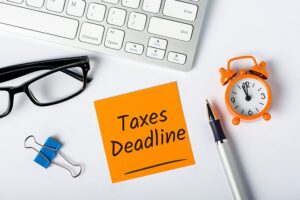 An orange sticky note with tax deadlines written in marker next to a laptop, glasses, pen, alarm clock, and paper clip in Springfield, IL.