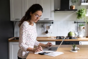 A young woman stands at the counter in her kitchen with paperwork, her laptop, and a calculator as she researches the best way to file her taxes in Springfield, IL.